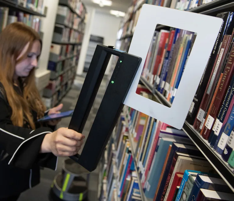 Une bibliothécaire lève l'antenne de récolement vers le haut d'un rayonnage